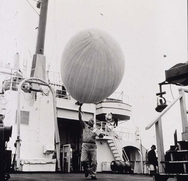 Old black and white picture. A guy is releasing a weather balloon, probably on a ship.